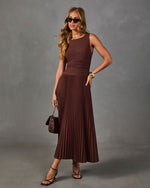 Woman in a brown sleeveless pleated midi dress with sunglasses, quilted brown handbag, gold cuff and strappy black heels