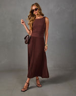 Woman wearing a brown sleeveless pleated midi dress with strappy heeled sandals, quilted brown bag, and cat-eye sunglasses