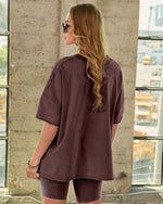 Woman wearing oversized mauve/burgundy t-shirt and matching bike shorts set, posed near industrial window, back view