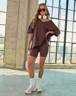 Woman wearing brown oversized v-neck top and matching biker shorts set with white sneakers, socks, and headphones around neck