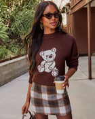 A woman wearing a brown knit sweater with a white teddy bear graphic and a brown, beige, and gray plaid mini skirt, holding a coffee cup in an outdoor walkway setting.