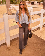 Woman in chambray ruffle top, dark wide-leg jeans, and western boots poses by white fence at horse ranch