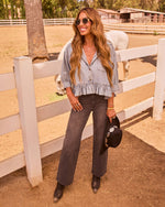 Woman in chambray ruffle top, dark wide-leg jeans, and western boots holding black cowboy hat at horse ranch fence