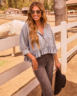Woman in cropped chambray ruffle top, dark flare jeans, gold sunglasses, holding black cowboy hat at ranch fence with white horse