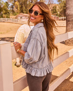 Woman in light-wash denim ruffle jacket and gray jeans smiling at horse ranch with white fence, wearing gold sunglasses