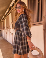 Woman in black plaid mini dress with lace bodice, gold jewelry, aviator sunglasses, holding woven bag at horse stable