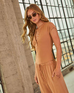 Woman wearing camel ribbed henley bodysuit and matching drawstring wide-leg pants with gold sunglasses, industrial backdrop
