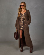 Woman in a long brown knit cardigan, floral mini slip dress, tall leather boots, sunglasses and a burgundy suede handbag