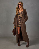 Woman in a long brown knit cardigan, floral mini slip dress, tall leather boots, sunglasses and a burgundy suede handbag