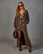 Woman in a long brown knit cardigan over a floral satin slip dress with tall brown boots, sunglasses and a burgundy handbag
