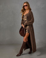 Woman in a long brown knit cardigan, floral satin slip dress, knee-high leather boots, and a suede hobo bag against gray backdrop