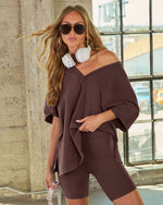 Woman wearing brown oversized V-neck sweatshirt and matching biker shorts set with white headphones and sunglasses