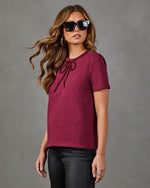 A woman modeling a short-sleeve crewneck t-shirt in deep burgundy with a decorative dark red bow detail at the neckline, paired with black pants against a textured gray studio background.