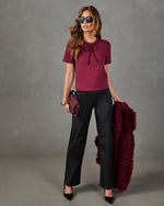 A woman models a short-sleeve burgundy knit top with a bow detail, paired with black wide-leg pants, holding a matching burgundy faux-fur jacket and a matching clutch against a gray studio backdrop.