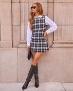 A woman models a black and white plaid pinafore mini dress over a white long-sleeve collared shirt, styled with black knee-high boots, sunglasses, and a small black handbag, posing against a beige stone wall.