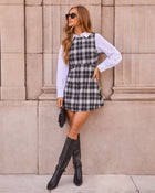 A woman models a black and white plaid pinafore mini dress over a white long-sleeve collared shirt, styled with black knee-high boots, sunglasses, and a small black handbag, posing against a beige stone wall.
