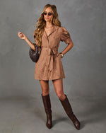 A woman models a brown mini dress with puff sleeves, a tie waist, and a subtle floral pattern, styled with dark brown knee-high boots, a dark brown textured handbag, and sunglasses against a gray studio backdrop.