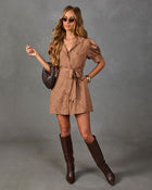 A woman models a brown mini dress with puff sleeves, a tie waist, and a subtle floral pattern, styled with dark brown knee-high boots, a dark brown textured handbag, and sunglasses against a gray studio backdrop.