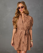 A woman modeling a brown short puff-sleeve dress with a tie waist and subtle tonal pattern, styled with sunglasses and gold jewelry against a textured gray studio background.