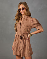 A woman modeling a brown short-sleeve mini dress with a tie waist and subtle tonal pattern, posed against a textured gray studio background.