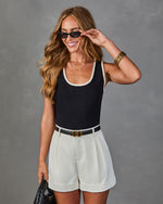 Woman wearing black knit bodysuit with cream trim, white pleated shorts, black belt, and small sunglasses, holding woven bag