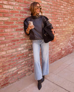 Woman in black leather jacket, wide-leg light wash jeans, black ankle boots and bag, leaning against brick wall holding phone