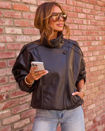 Woman in oversized dark brown leather moto jacket, aviator sunglasses, light jeans, holding smartphone against brick wall