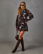 Brown long-sleeve tiered mini dress with blue floral print, styled with dark suede knee-high boots and a woven shoulder bag