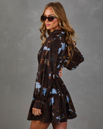 Woman in a brown long-sleeve mini dress with blue floral print, tiered skirt and cat-eye sunglasses against a gray backdrop