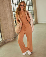 Wide-leg caramel drawstring sweatpants with side pockets, paired with white sneakers, worn by a woman in an industrial setting.