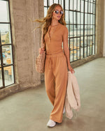 Wide-leg caramel drawstring sweatpants with side pockets, paired with white sneakers, worn by a woman in an industrial setting.