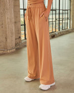 Wide-leg caramel drawstring sweatpants with side pockets, paired with white sneakers, worn by a woman in an industrial setting.