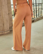 Wide-leg caramel drawstring sweatpants with side pockets, paired with white sneakers, worn by a woman in an industrial setting.