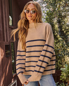 A woman wearing a beige knit sweater with navy blue horizontal stripes, styled with light blue jeans and round sunglasses, standing outdoors in front of a wooden house and green foliage.