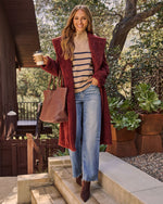 A woman stands on outdoor stone steps wearing a burgundy fuzzy open-front coat over a striped sweater, blue jeans, and brown ankle boots, while carrying a brown woven tote bag and holding a coffee cup.