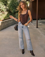 A woman stands outdoors wearing a brown ruffled camisole top, light-wash distressed wide-leg jeans, and dark brown ankle boots, with a small dark shoulder bag.