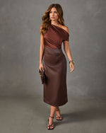 Woman in a brown satin one-shoulder top and faux leather midi skirt with strappy heels, gold jewelry, and a suede clutch