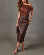 Woman in brown satin asymmetric off-shoulder top and faux leather midi skirt with strappy heeled sandals and gold jewelry