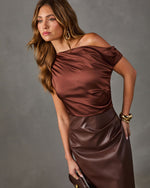 Woman in a chocolate brown satin asymmetrical off-shoulder top and faux leather midi skirt with gold cuff bracelet and earrings