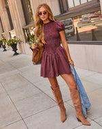 A woman stands on a city sidewalk wearing a short-sleeve burgundy and black checkered mini dress with a peplum waist, paired with tan knee-high boots and holding a blue denim jacket and a brown shoulder bag.