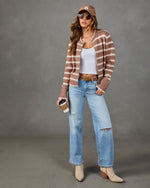 A woman models a brown and beige striped knit cardigan, styled with a white tank top, light-wash distressed jeans, beige ankle boots, a brown baseball cap, and sunglasses, holding a coffee cup against a gray studio backdrop.