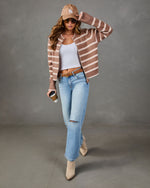 A woman models a brown and beige striped knit cardigan, styled with a white tank top, light-wash distressed jeans, beige ankle boots, a tan baseball cap, and sunglasses against a gray studio backdrop.