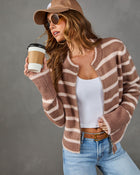 A woman wearing a brown and beige striped fuzzy zip-up cardigan, a white tank top, blue jeans, a brown cap, and sunglasses, holding a coffee cup against a gray studio background.