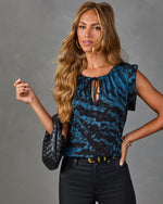 Woman wearing blue black abstract print tie-neck blouse, black skinny jeans, gold belt, holding woven leather bag