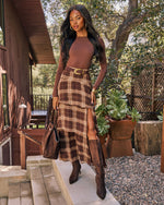 A woman stands on outdoor steps in front of a wooden house, wearing a brown long-sleeve bodysuit, a brown and tan plaid midi skirt with a high slit and gold belt, brown knee-high boots, and carrying a large brown tote bag.