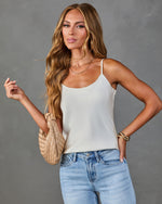 Woman wearing white satin cami tank top and light wash high-waist jeans, holding woven tan mini bag, layered gold necklaces