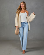 Woman wearing oversized cream knit cardigan, white cami, light wash wide-leg jeans, nude heels, holding woven clutch bag