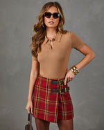 A woman modeling a tan short-sleeve knit polo top and a red plaid mini skirt with double black belts featuring gold buckles, styled with gold bracelets and sunglasses against a gray studio background.
