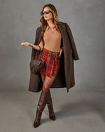 A woman models a brown longline coat over a tan top, a red and yellow plaid mini skirt, brown knee-high boots, and a brown woven handbag against a gray studio backdrop.
