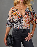 Woman wearing multicolor ikat print button-front blouse with puff sleeves, dark jeans, gold layered necklaces, black woven bag
